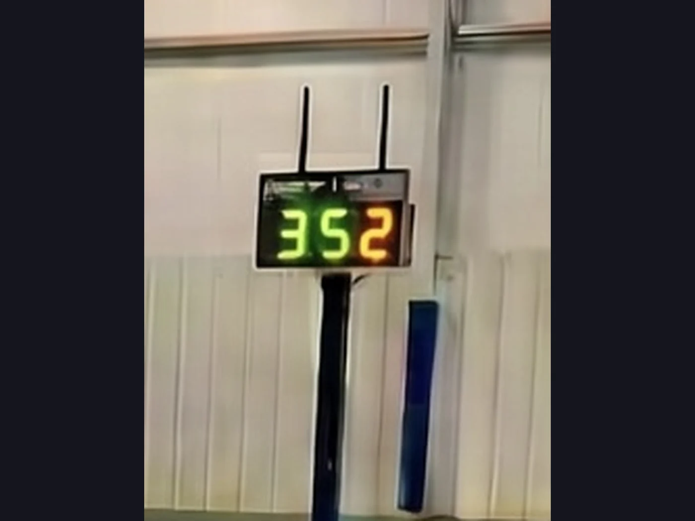 Eye mounted on a net-post pole at an indoor pickleball court, its bright LED scoreboard showing the score 3-5-2 in green and amber